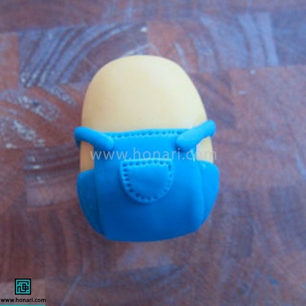 آموزش عروسک خمیری مینیون-مرحله 10 نیم دایره کوچکی درست کرده ،آنرا به عنوان جیب در جلوی شلوار وصل میکنیم . در صورت دلخواه میتوانید با استفاده از سوزن طرح جای دوخت روی آن ایجاد کنید